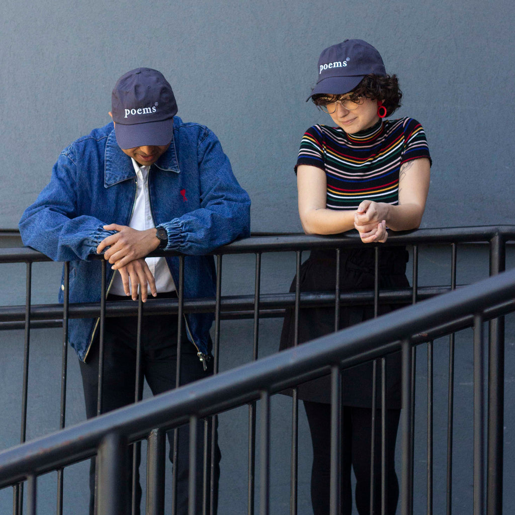  Photo: Male and female models wearing navy blue ball caps with 'poems' embroidered on them. The models are leaning casually against a black metal railing. 