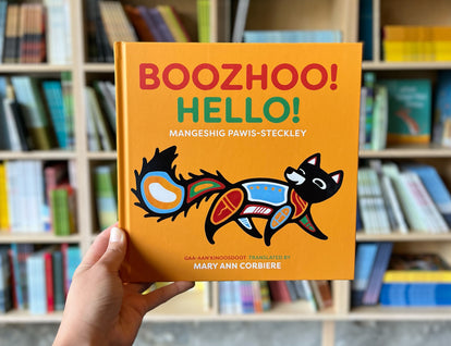 A photograph of a hand holding up a copy of Boozhoo!/Hello! against a background of bookshelves.