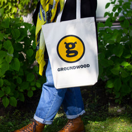  Photo: A white tote bag printed with the iconic encircled 'G' of the Groundwood Books logo. The bag is being modeled by someone standing in a grassy outdoor area. 