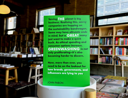 Photo of The Great Greenwashing by John Pabon. A bright green cover filled with two paragraphs of bold black and white text. The book is upright on a plant stand, and the House of Anansi bookshop is in the background.  
