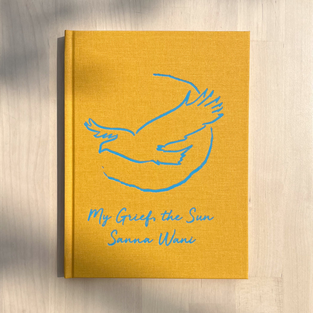  A photo of the book My Grief, the Sun on a wooden table. The book is yellow hardcover edition. A bird with a quarter-complete circle behind it is drawn on the cover in blue, and the title is in blue cursive below the image.  