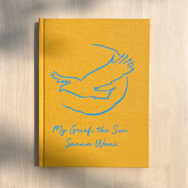  A photo of the book My Grief, the Sun on a wooden table. The book is yellow hardcover edition. A bird with a quarter-complete circle behind it is drawn on the cover in blue, and the title is in blue cursive below the image.  