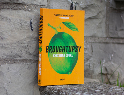 Photo of Broughtupsy by Christina Cooke. A bright orange cover with a large illustration of a green lime in the center. A quote in white text that reads: “A Writer of Immense Heart” - Canisia Lubrin. The book is leaning against a textured stone wall.