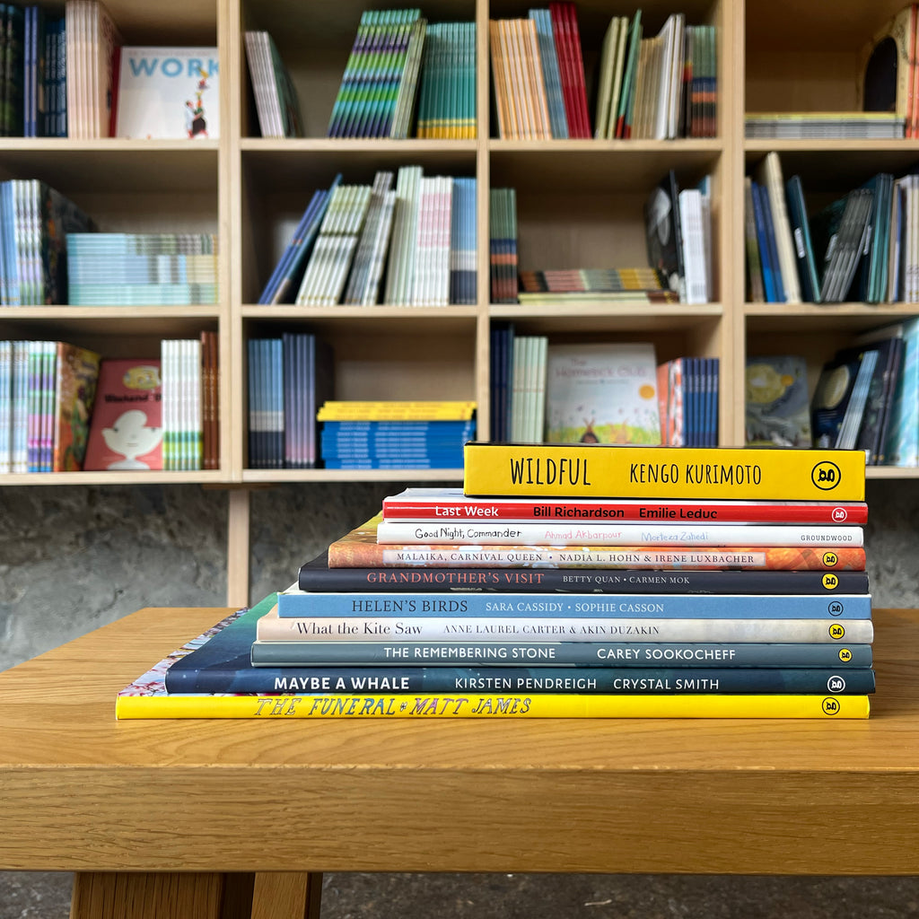  Photo of books featured in the Navigating Death, Loss, Grief bundle. The books are stacked on a wooden stool, with bookshelves in the background. The spines face out. 