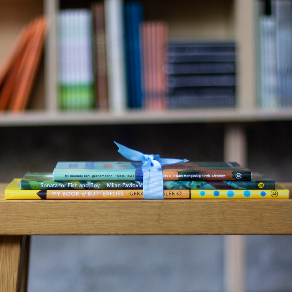  A photo of a stack of picture books, spines out, sitton on a wooden bench. They are tied together with a blue ribbon. The books are: Mii maanda ezhi-gkendmaanh / This Is How I Know, Sonata for Fish and Boy, and My Book of Butterflies. 