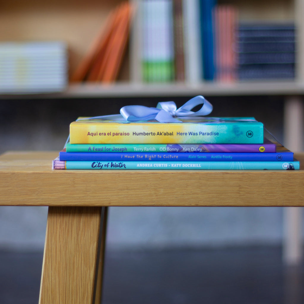  A photo of four books in a stack on a wooden bench, tied together with narrow satin blue ribbon. The books are: Aquí era el paraíso / Here Was Paradise, A Feast for Joseph, I Have the Right to Culture, and City of Water. 