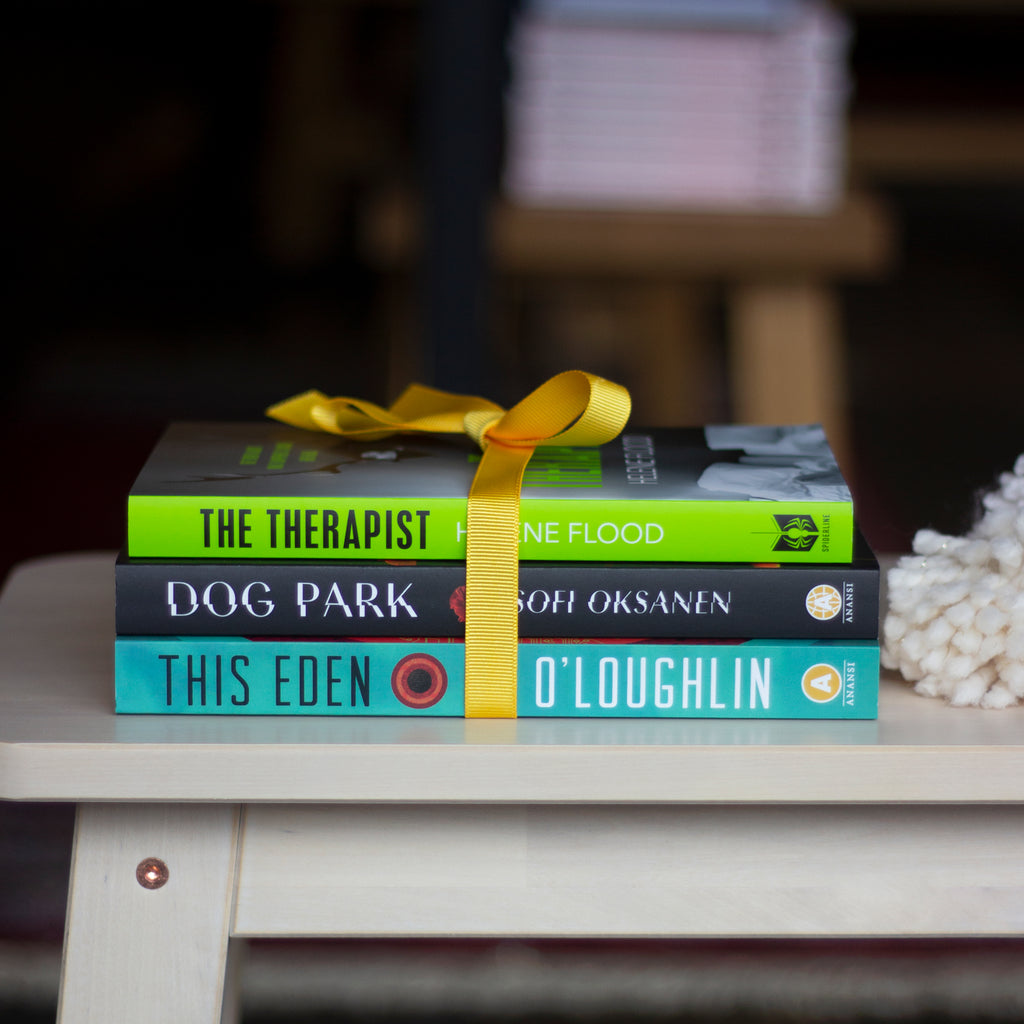  A photo of copies of The Therapist, Dog Park, and This Eden sitting on a bench tied together with a bright yellow ribbon. Soft, yarn, off-white pom-poms also sit atop the bench next to the stack. 