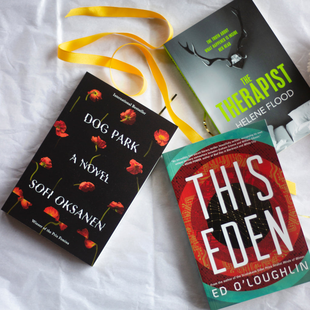  A photo of copies of The Therapist, Dog Park, and This Eden laying flat on top of white tissue paper. A bright yellow ribbon snakes around them. The cover of Dog Park features red poppies on a black background, intermittently sliced in half and shifted. The cover of This Eden features concentric circles, each containing different tech-y illustrations. The cover of The Therapist features a large mounted set of deer horns above two twin beds with white sheets pushed together. 