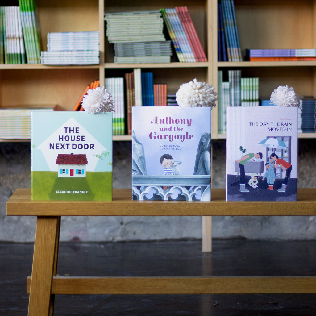  A photo of three picture books sitting upright on a wooden bench. The books are: The House Next Door, Anthony and the Gargoyle, and The Day the Rain Moved In.  