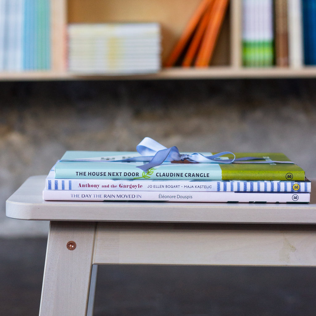  A photo of a stack of picture books, spines-out, sitting on a wooden bench. They are tied together with a narrow, satin, blue ribbon. 
