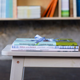  A photo of a stack of picture books, spines-out, sitting on a wooden bench. They are tied together with a narrow, satin, blue ribbon. 