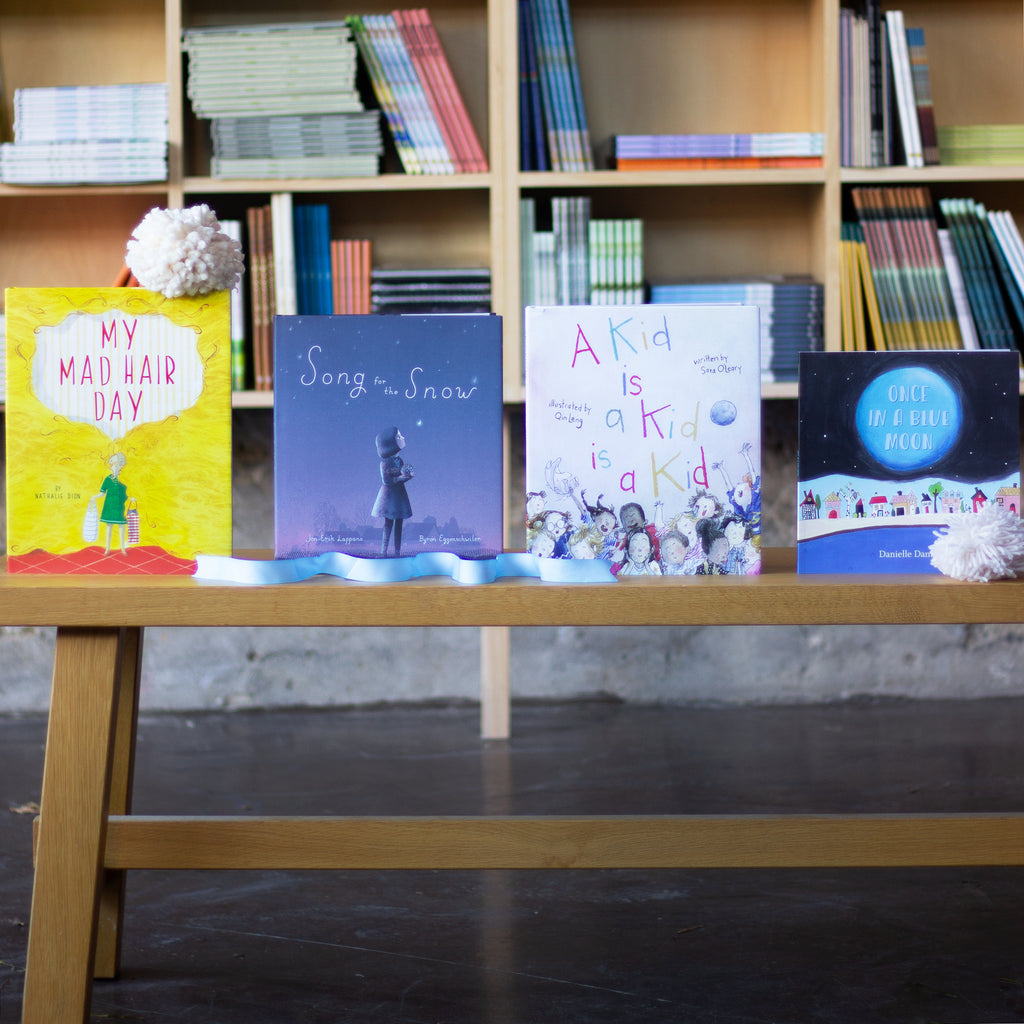  A photo of four picture books sitting upright top of a wooden bench. An off-white pompom made of thick yarn sits on top of one of the books and below another. The books are: Once in a Blue Mono, Song for the Snow, A Kid Is a Kid Is a Kid, and My Mad Hair Day. 