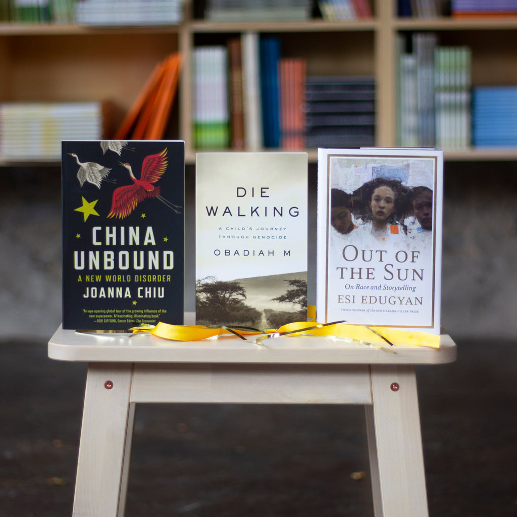  A photo of copies of China Unbound, Die Walking, and Out of the Sun sitting upright on a wooden bench. Also on top of the bench is a scattering of golden tinsel and a bright yellow ribbon. The cover of China Unbound features silver and red cranes flying upward, surrounded by yellow stars. The cover of Die Walking features a sepia photograph of dry countryside, with a path cutting through low scrubby trees and shrubs. The cover of Out of the Sun features a painting of three black women facing the viewer. 