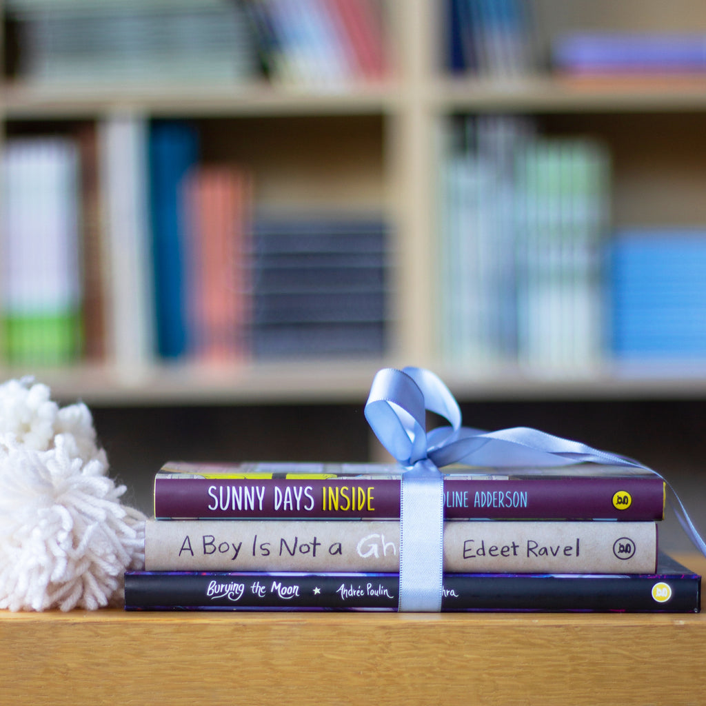  A photo of three books in a stack, spines out, on top of a wooden bench. A pompom made of off-white, thick yarn sits next to the stack. The books include: Burying the Moon, A Boy Is Not a Ghost, and Sunny Days Inside. 