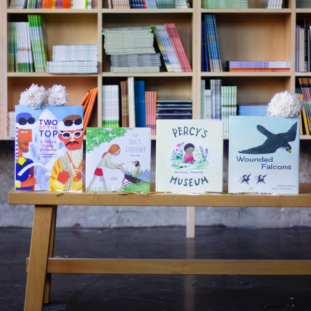  A photo of four picture books sitting upright on a wooden bench. Off-white pompoms made of thick yarn sit on top of the books. The books are: The Dog’s Gardener, Percy’s Museum, Wounded Falcons, and Two at the Top. 