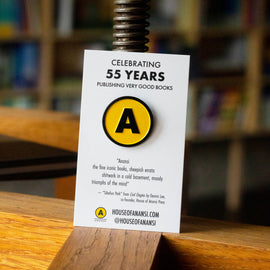  A photo of an enamel pin on a business card on a wooden shelf in the Anansi bookshop. The pin is the Anansi logo - a yellow circle, outlined in black, with a black, sans serif A in the middle. On the card is text that says Celebrating 55 years publishing very good books. A poem: 